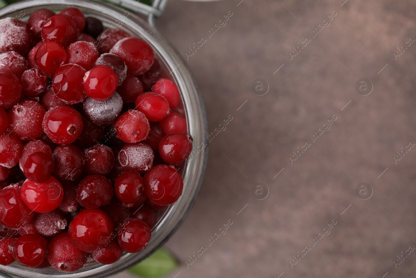 Photo of Frozen red cranberries in glass jar on brown textured table, top view. Space for text