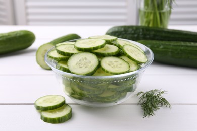 Photo of Cut cucumber in glass bowl, fresh vegetables and dill on white wooden table, closeup