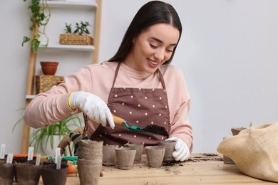 Photo of Young woman adding soil into peat pots at wooden table indoors. Growing vegetable seeds