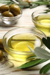 Photo of Bowls with cooking oil, olives and green leaves on white wooden table, closeup