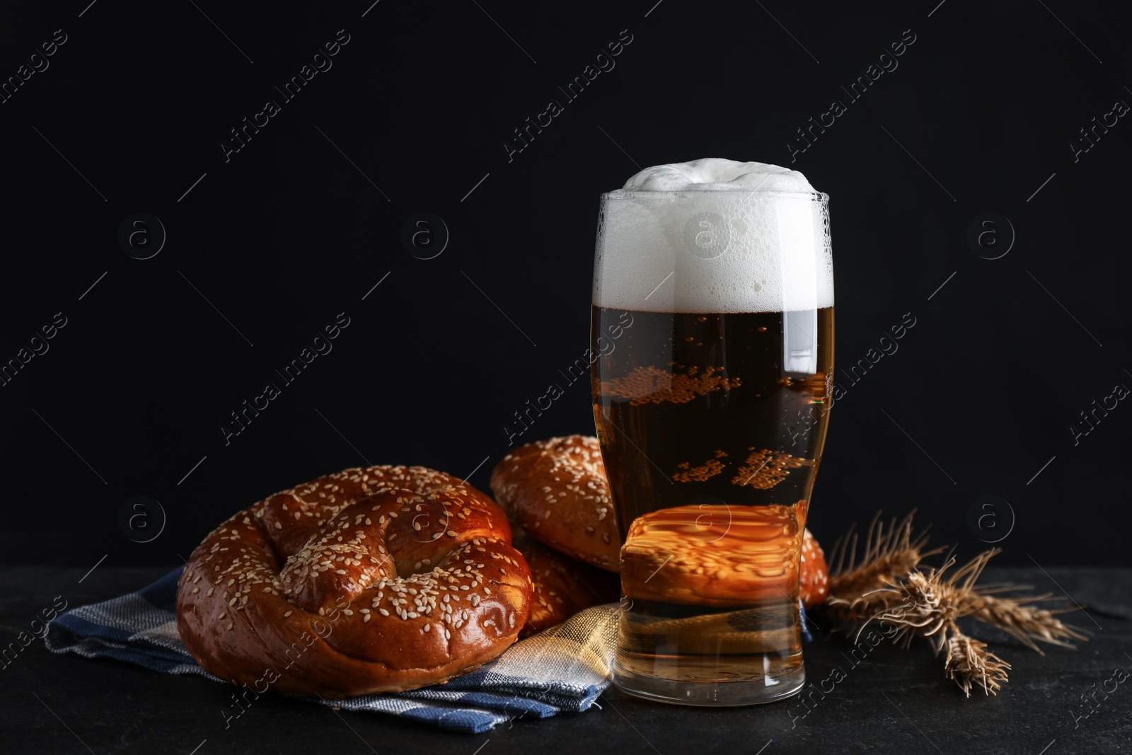 Photo of Tasty pretzels, glass of beer and wheat spikes on black table against dark background