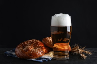 Tasty pretzels, glass of beer and wheat spikes on black table against dark background