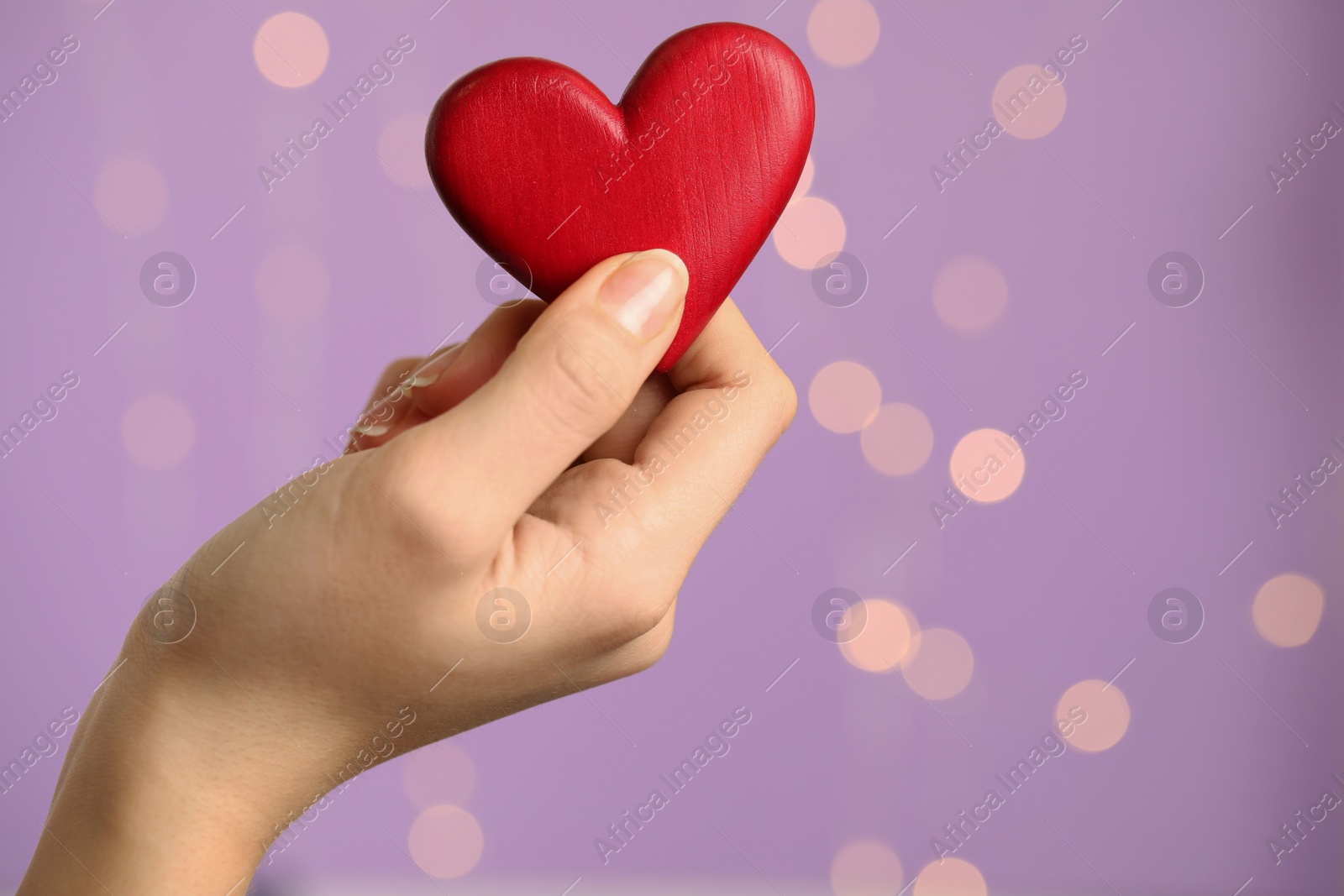 Photo of Closeup view of woman holding red heart on violet background with festive lights, space for text. St. Valentine's day