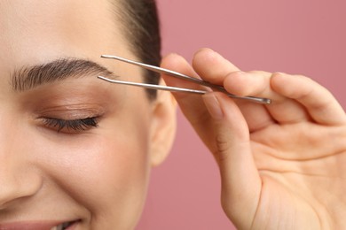 Photo of Eyebrow correction. Young woman with tweezers on pink background, closeup