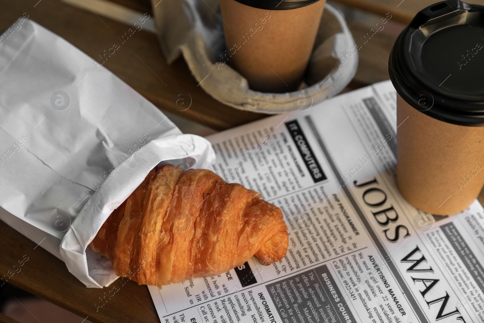Photo of Tasty croissant, newspaper and paper cups of coffee on wooden table, closeup