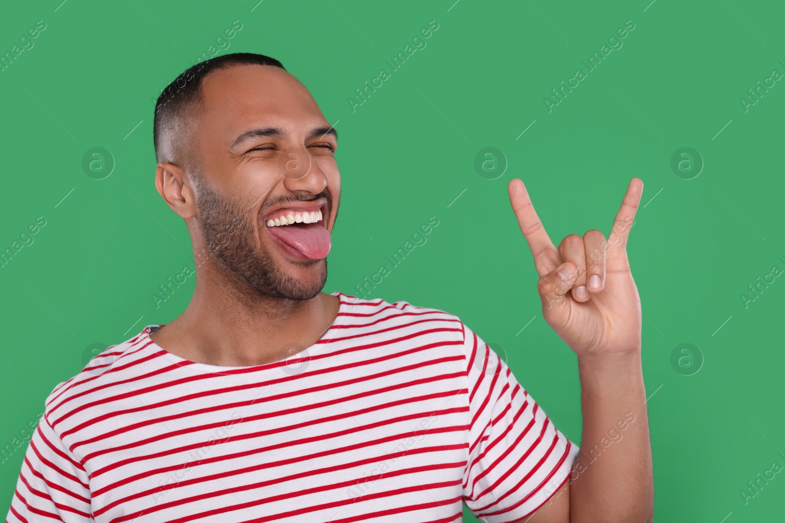 Photo of Happy young man showing his tongue and rock gesture on green background