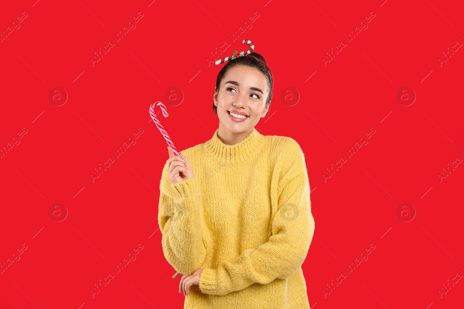 Photo of Young woman in yellow sweater and festive headband holding candy cane on red background