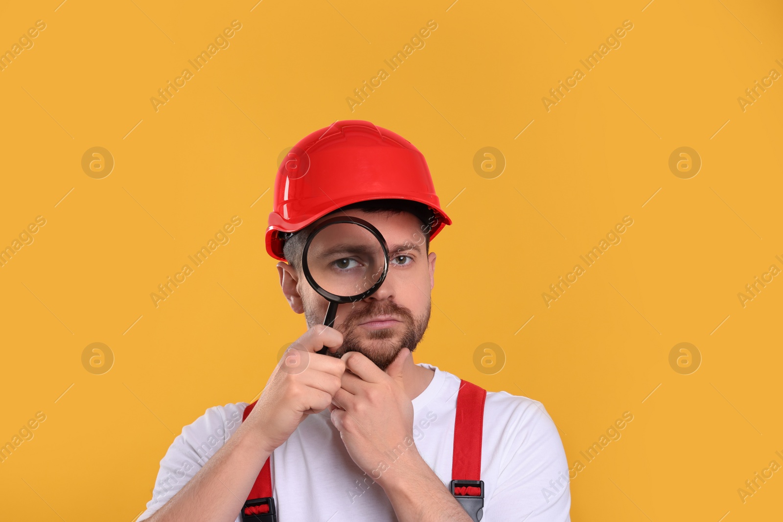 Photo of Confused builder looking through magnifier glass on yellow background