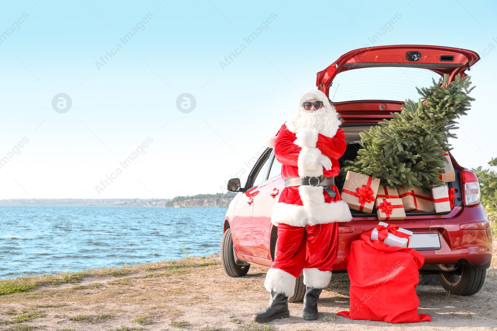 Photo of Authentic Santa Claus near red car with gift boxes and Christmas tree on beach