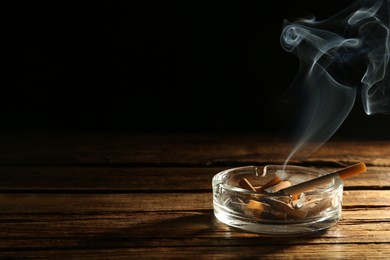 Photo of Smoldering cigarette in glass ashtray on wooden table against black background. Space for text