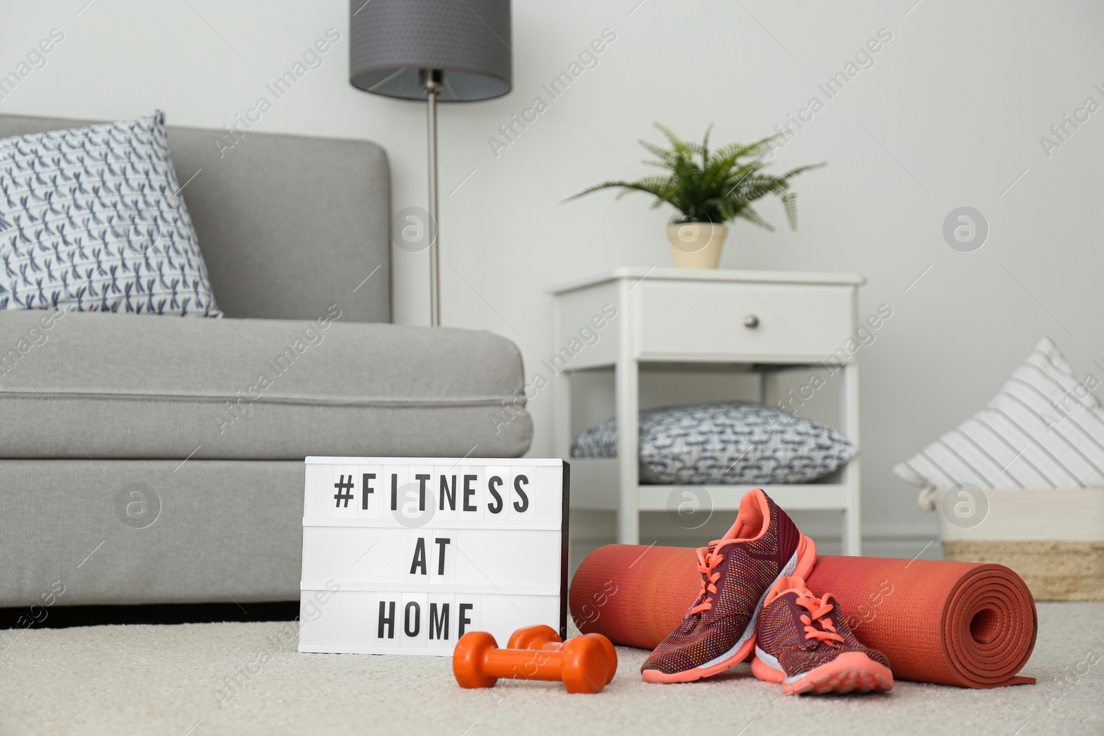 Photo of Sport equipment and lightbox with hashtag FITNESS AT HOME on floor indoors. Message to promote self-isolation during COVID‑19 pandemic