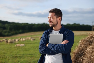 Photo of Smiling farmer with tablet near hay bale on pasture. Space for text