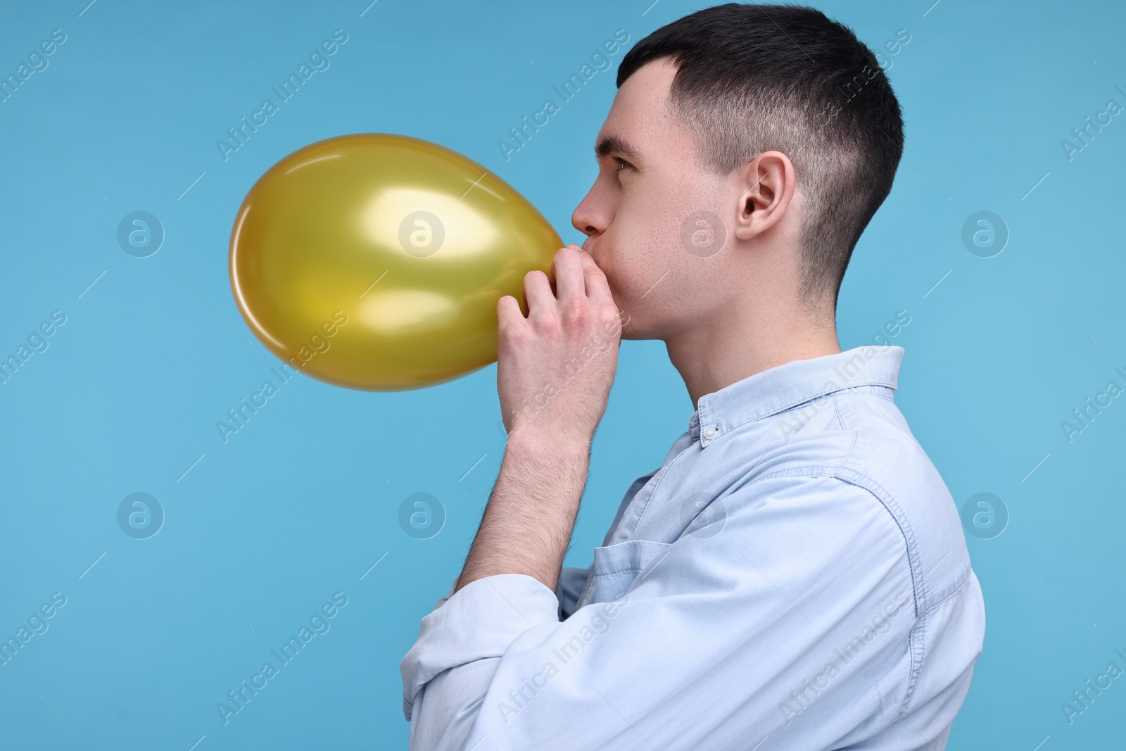 Photo of Young man inflating golden balloon on light blue background