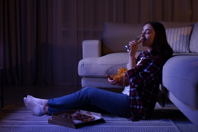 Photo of Young woman drinking from tin can and eating chips while watching TV in room at night. Bad habit