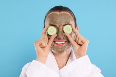 Photo of Beautiful woman with clay mask covering eyes with pieces of cucumber on light blue background