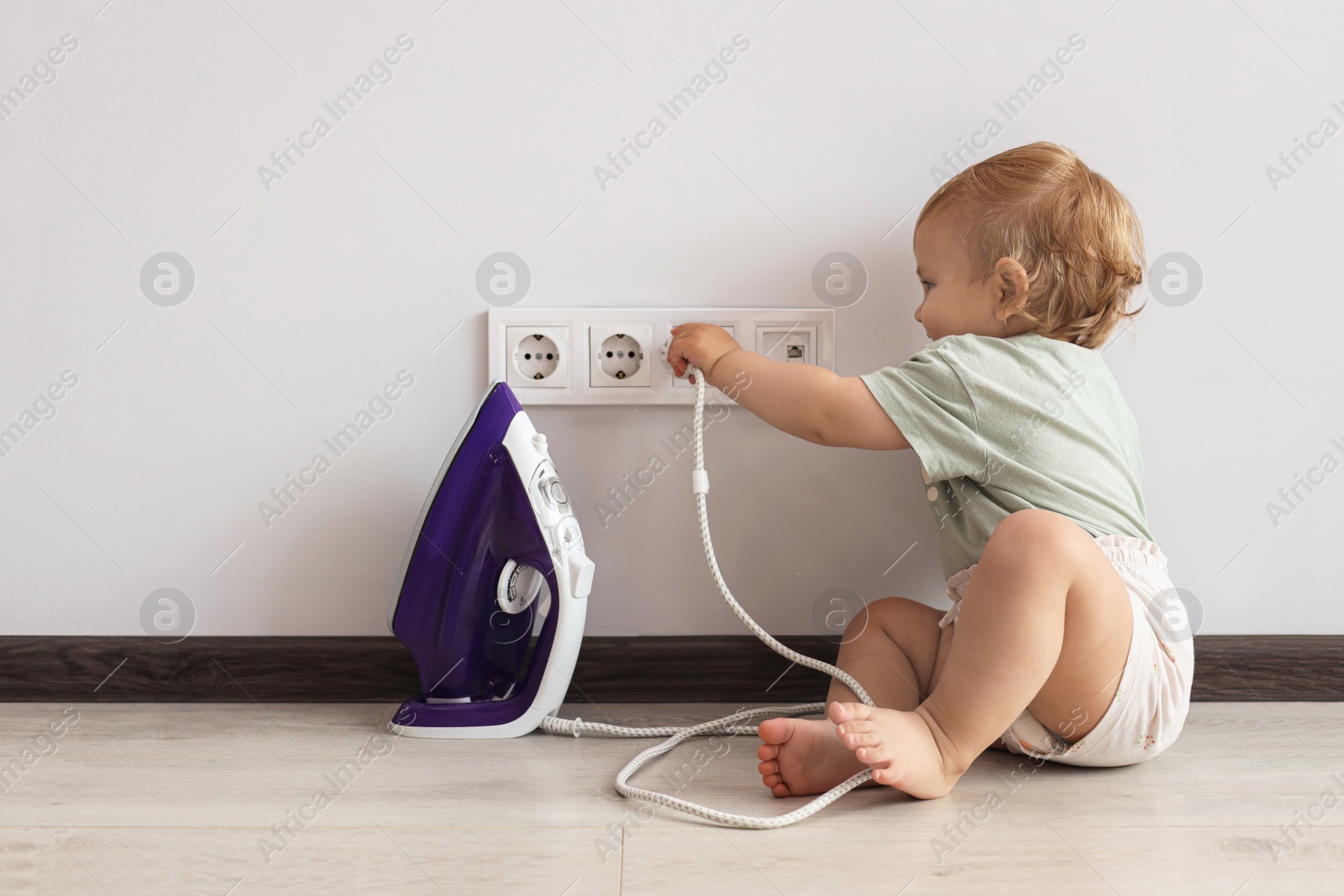 Photo of Cute baby playing with electrical socket and iron plug at home. Dangerous situation