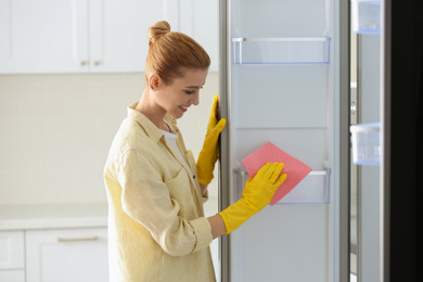 Photo of Woman in rubber gloves cleaning empty refrigerator at home