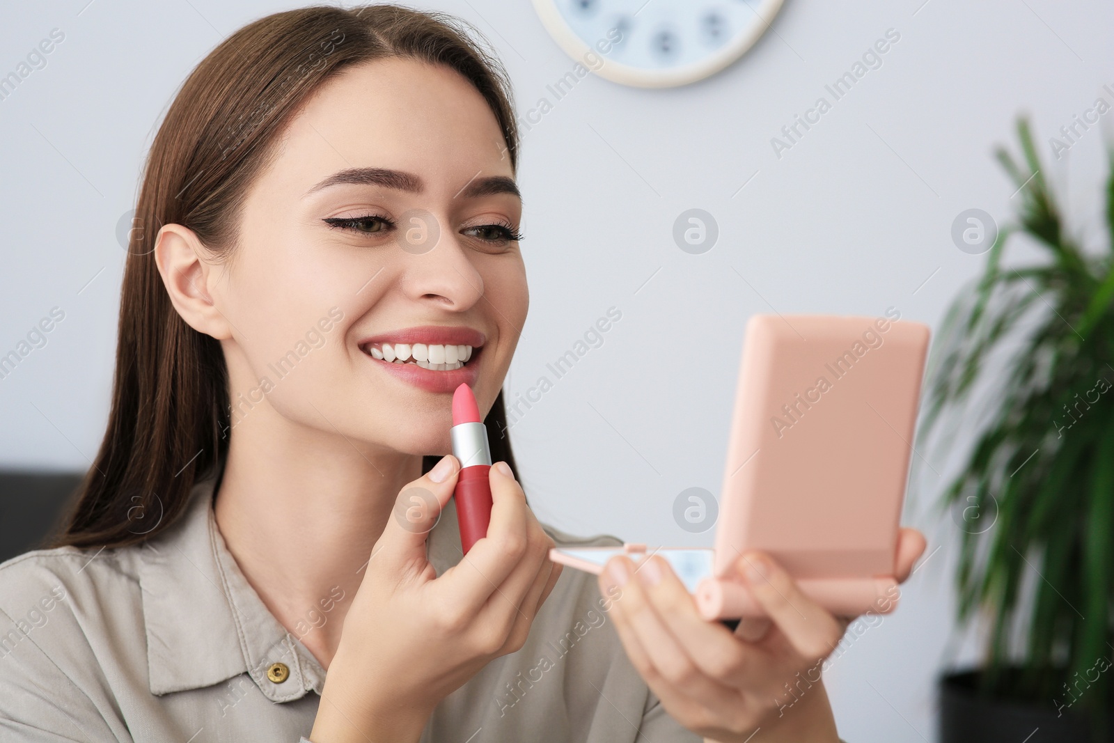 Photo of Young woman with cosmetic pocket mirror applying lipstick indoors