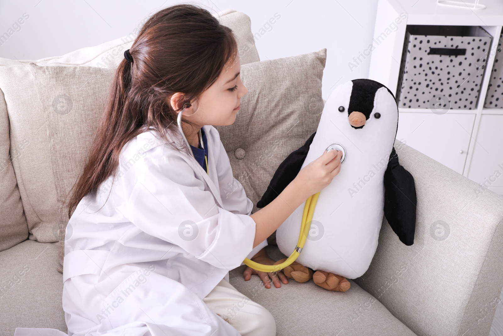 Photo of Little girl playing doctor with toy penguin on sofa indoors. Pediatrician practice