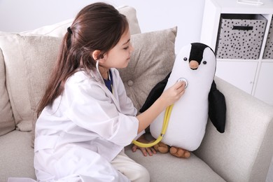 Photo of Little girl playing doctor with toy penguin on sofa indoors. Pediatrician practice