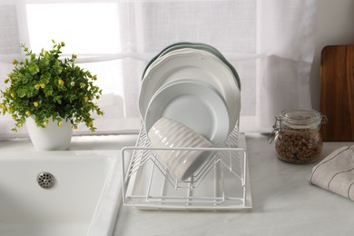 Photo of Drainer with different clean dishware, cup and houseplant near sink on light table indoors