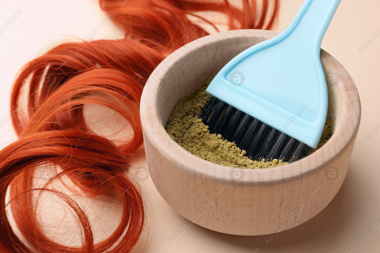 Photo of Bowl of henna powder, brush and red strand on beige background, closeup. Natural hair coloring