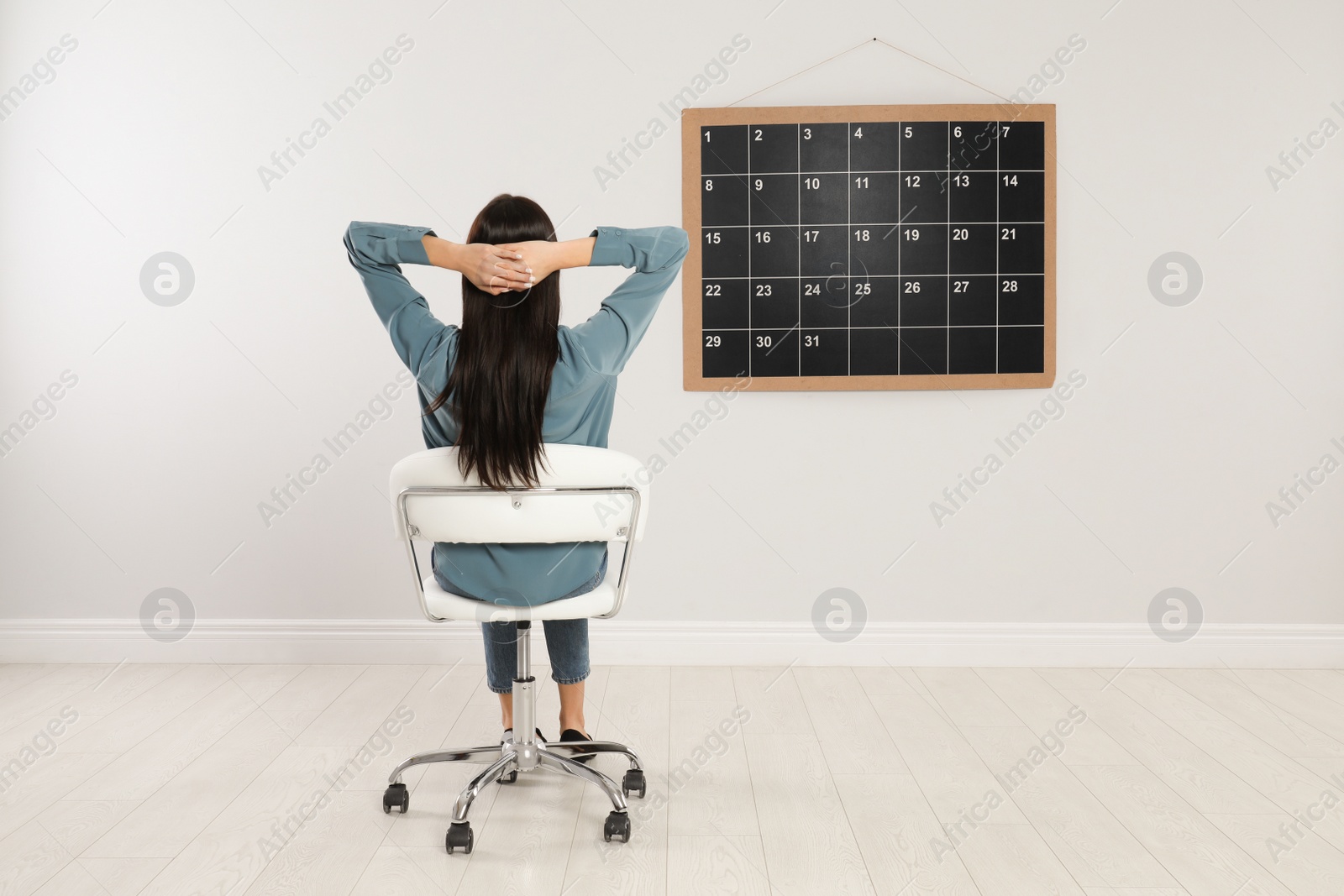 Photo of Young woman sitting near board calendar indoors, back view