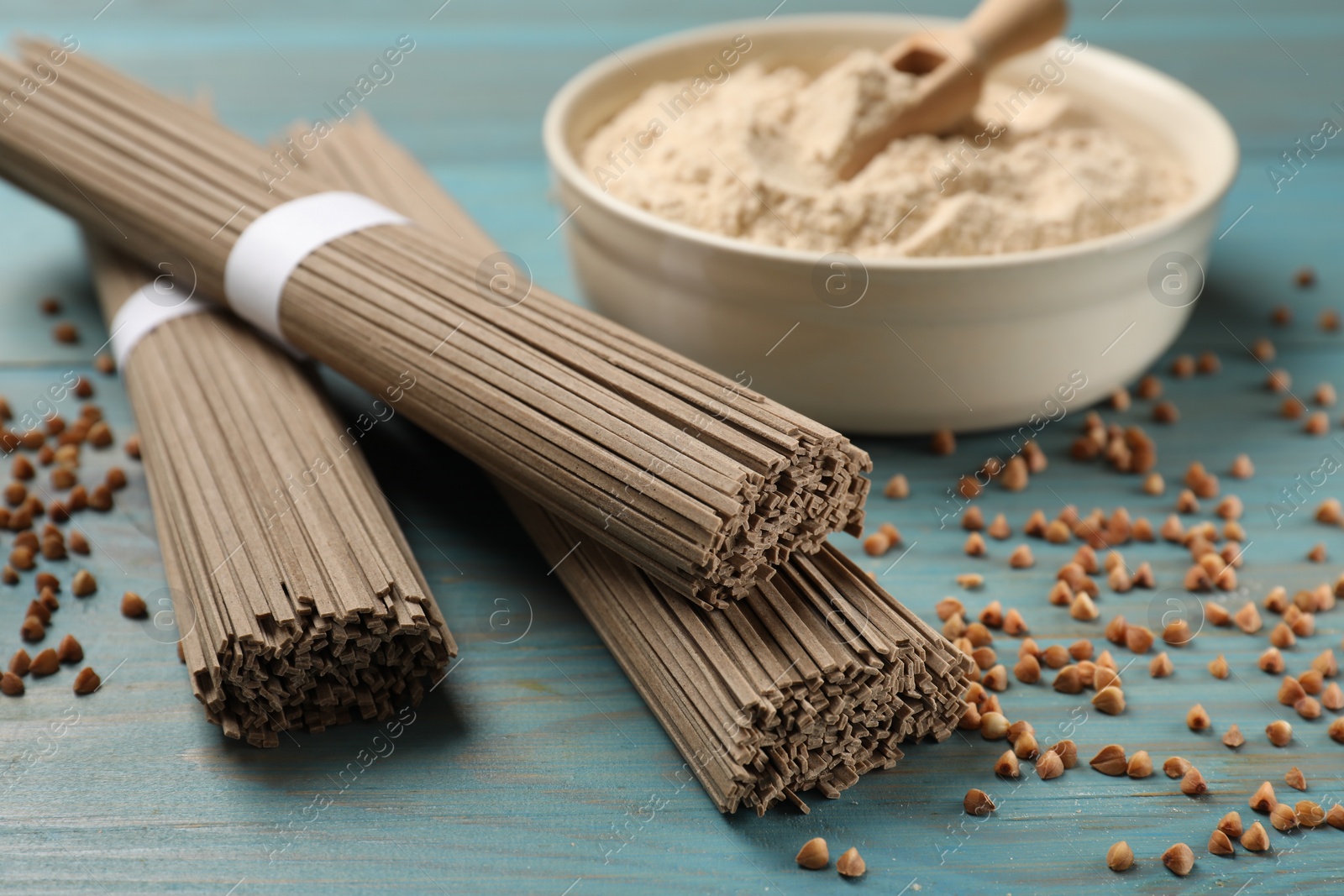 Photo of Uncooked buckwheat noodles (soba), flour and grains on light blue wooden table, closeup