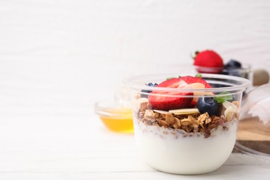 Photo of Tasty granola with berries, yogurt and almond flakes in plastic cup on white wooden table. Space for text