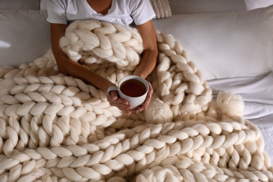 Photo of Woman covered with knitted plaid holding cup of tea in bed, closeup