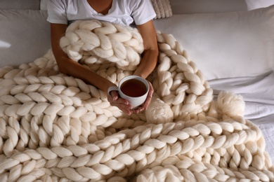 Woman covered with knitted plaid holding cup of tea in bed, closeup