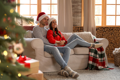 Happy young couple wearing Santa hats on sofa in living room decorated for Christmas