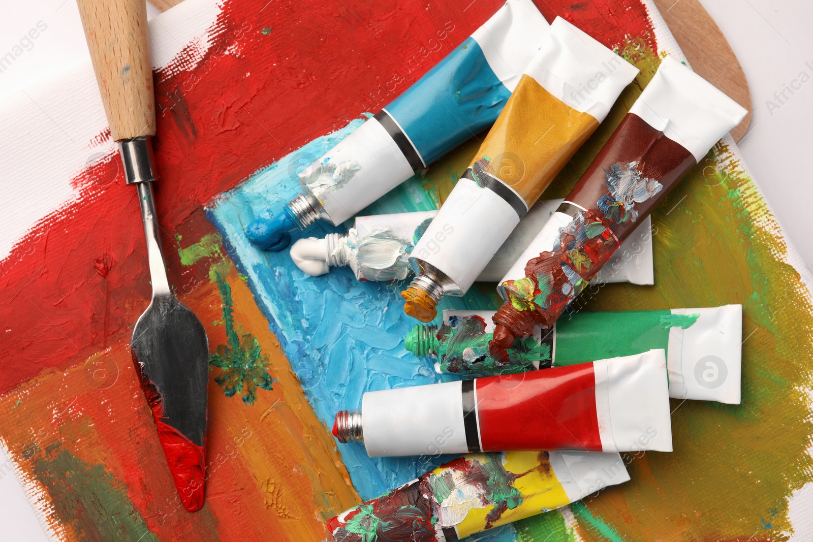 Photo of Tubes of colorful oil paints, spatula and canvas with abstract painting on white table, top view