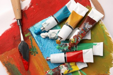 Photo of Tubes of colorful oil paints, spatula and canvas with abstract painting on white table, top view
