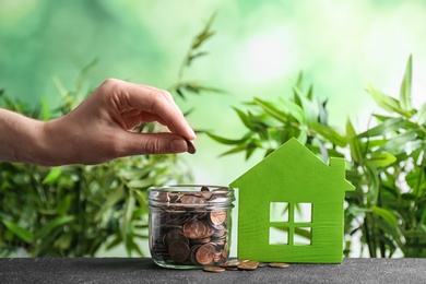 Woman putting coin into jar near house model on table against blurred background. Space for text