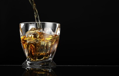 Pouring whiskey into glass with ice cubes at table against black background, space for text