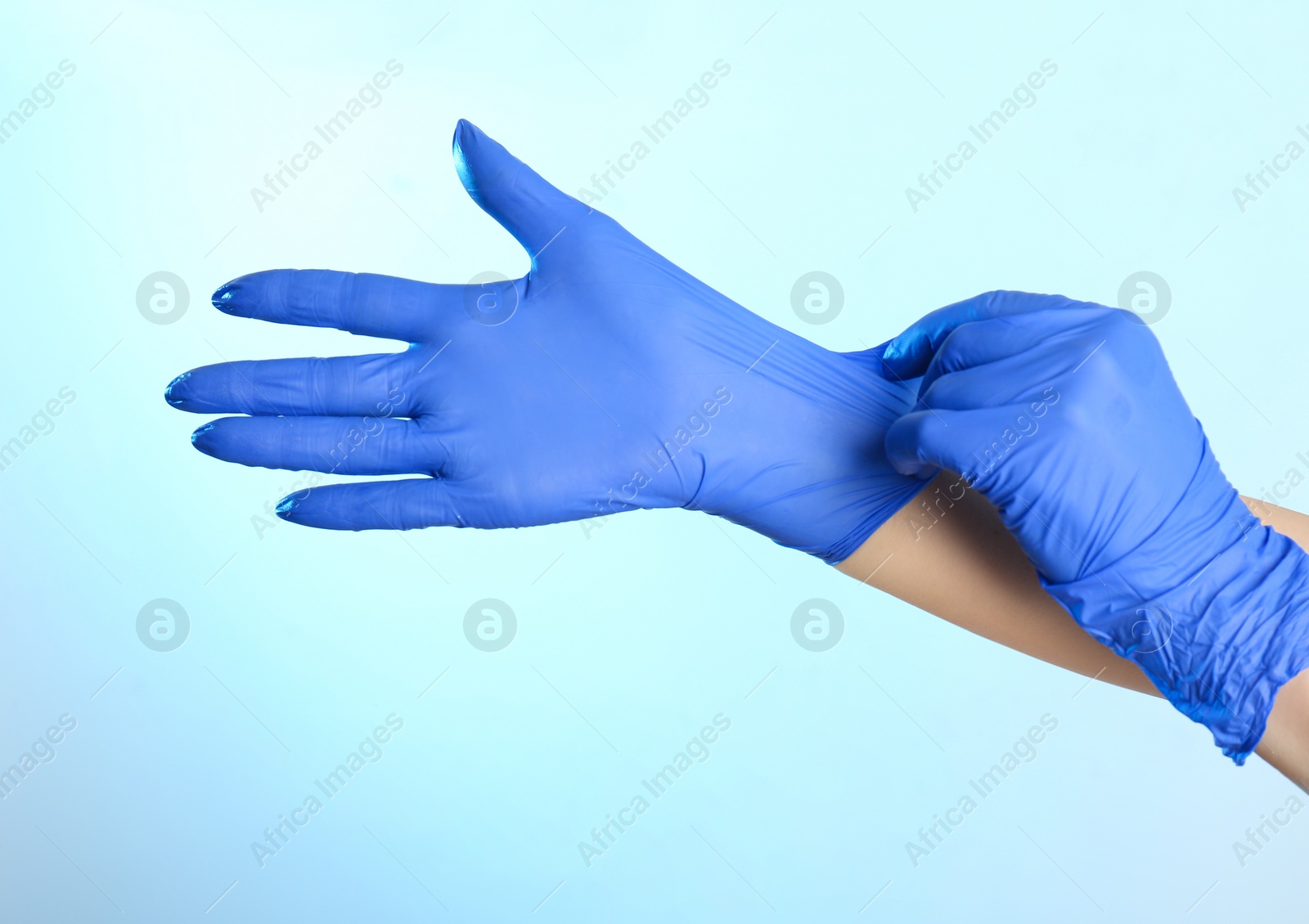 Photo of Woman putting on latex gloves against light blue background, closeup of hands