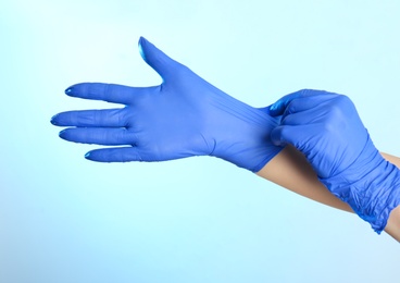 Photo of Woman putting on latex gloves against light blue background, closeup of hands