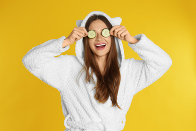 Young woman in bathrobe with cucumber slices on yellow background