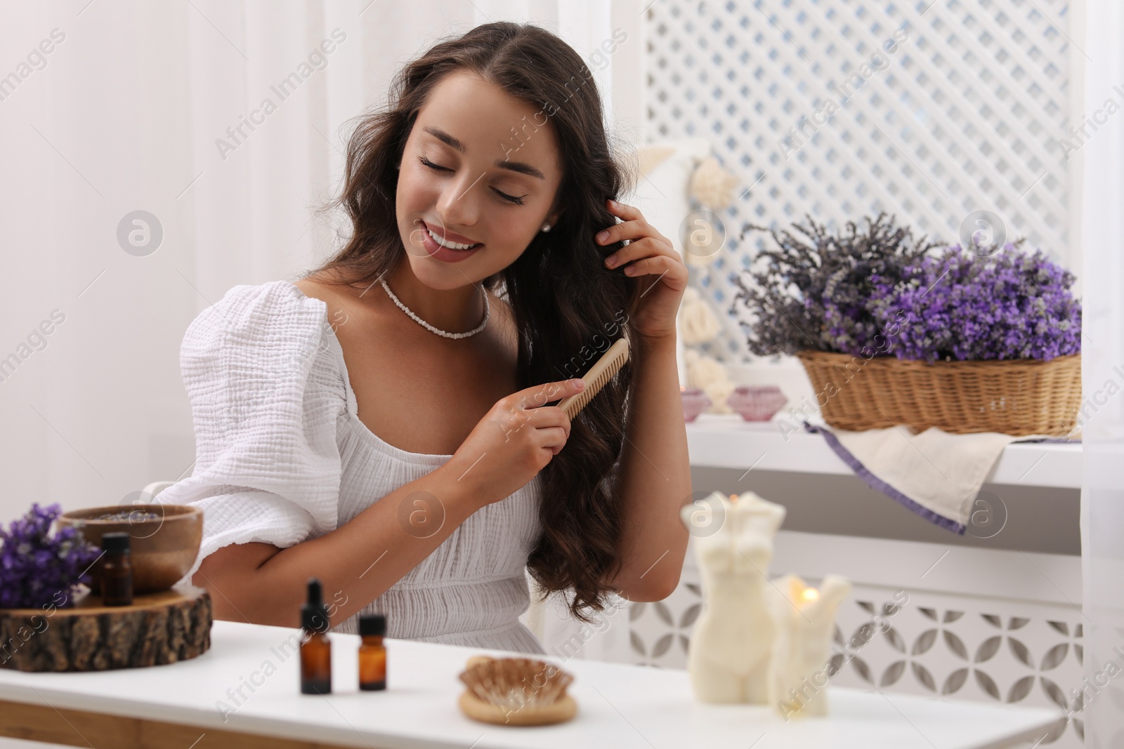 Photo of Beautiful young woman combing hair after applying essential oil at table indoors