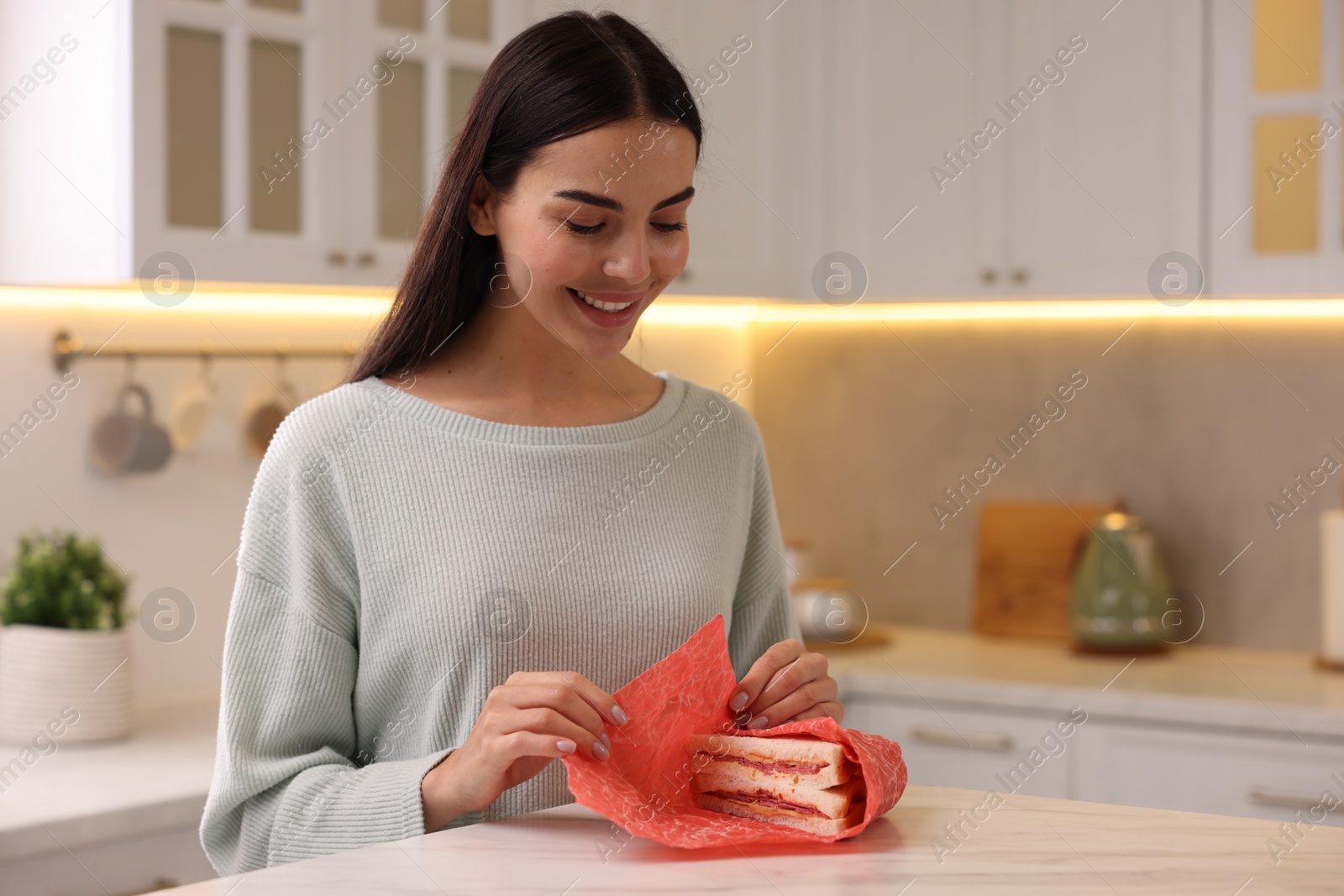 Photo of Happy woman packing sandwich into beeswax food wrap at table in kitchen