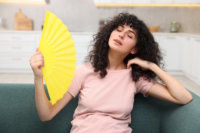 Photo of Young woman waving yellow hand fan to cool herself on sofa at home