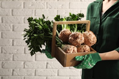Woman holding wooden crate with raw celery roots near white brick wall, closeup