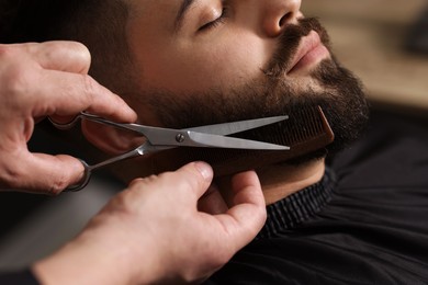 Photo of Professional barber trimming client's beard with scissors in barbershop, closeup