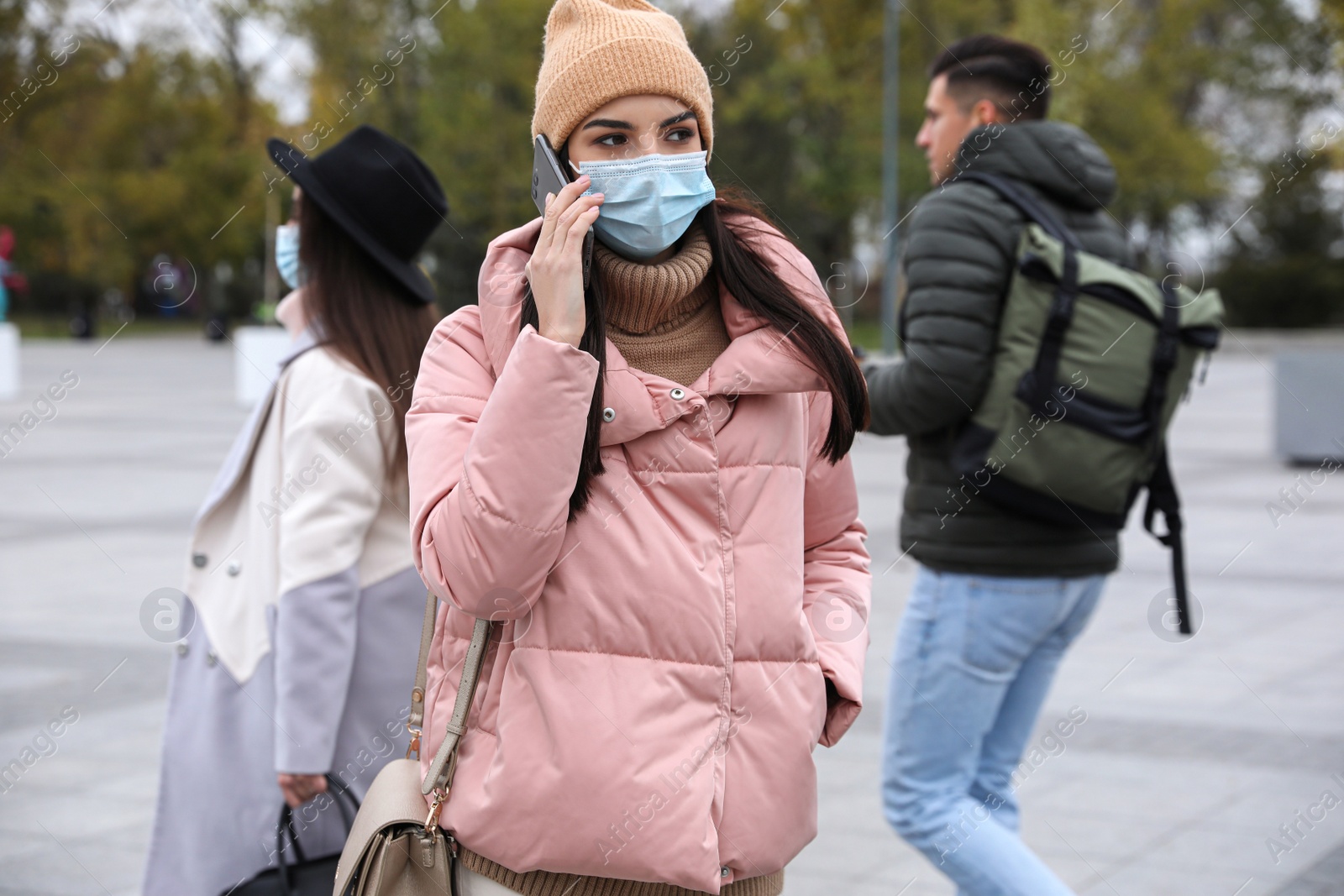 Photo of Young woman in medical face mask talking on phone while walking outdoors. Personal protection during COVID-19 pandemic