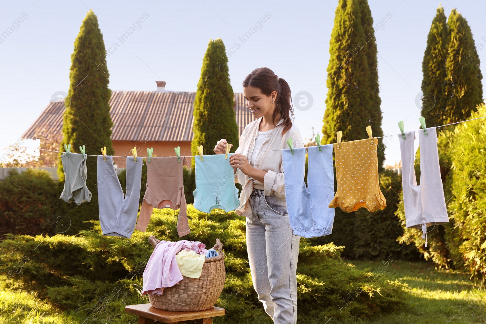 Photo of Smiling woman hanging baby clothes with clothespins on washing line for drying in backyard