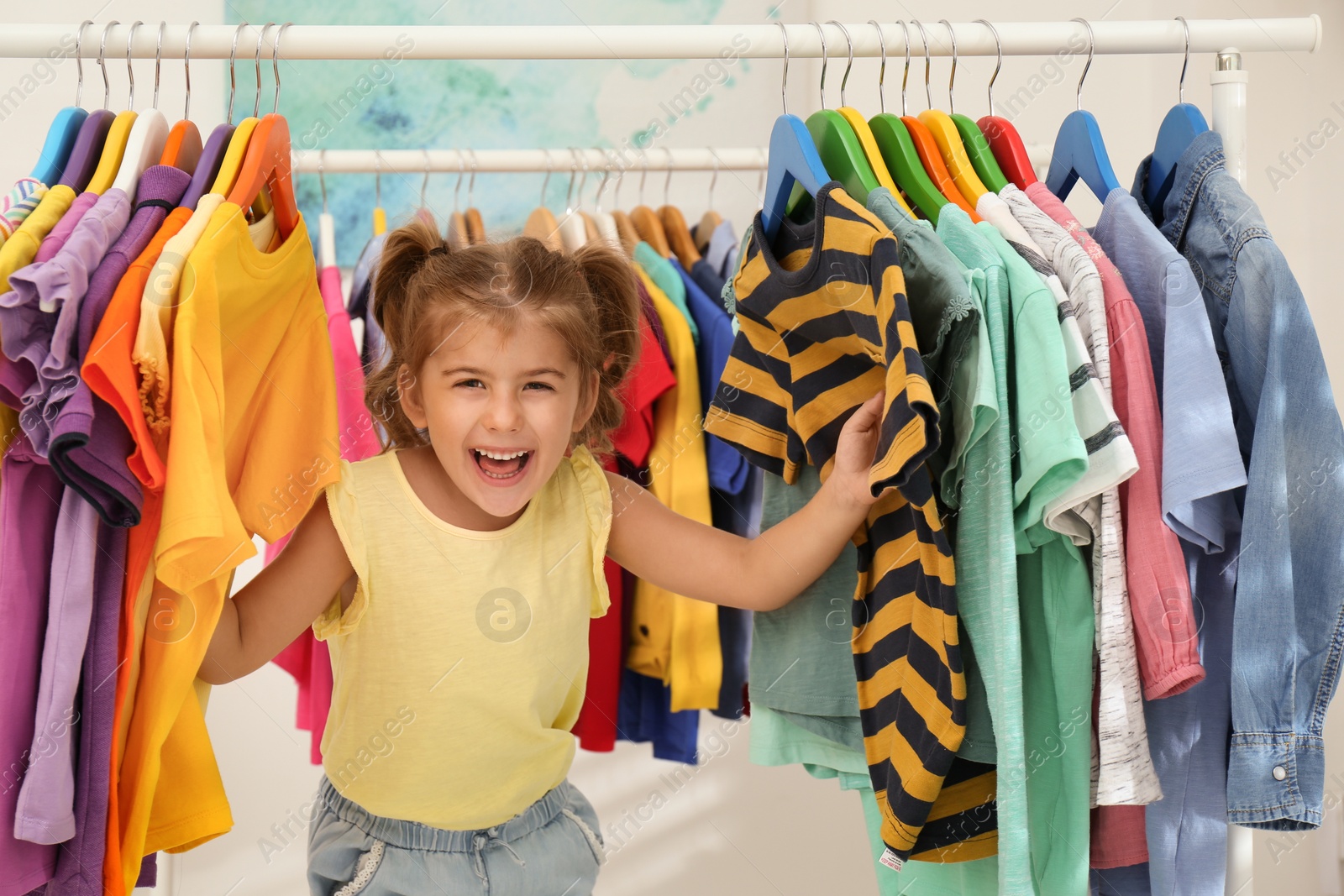 Photo of Funny little girl choosing clothes on rack indoors