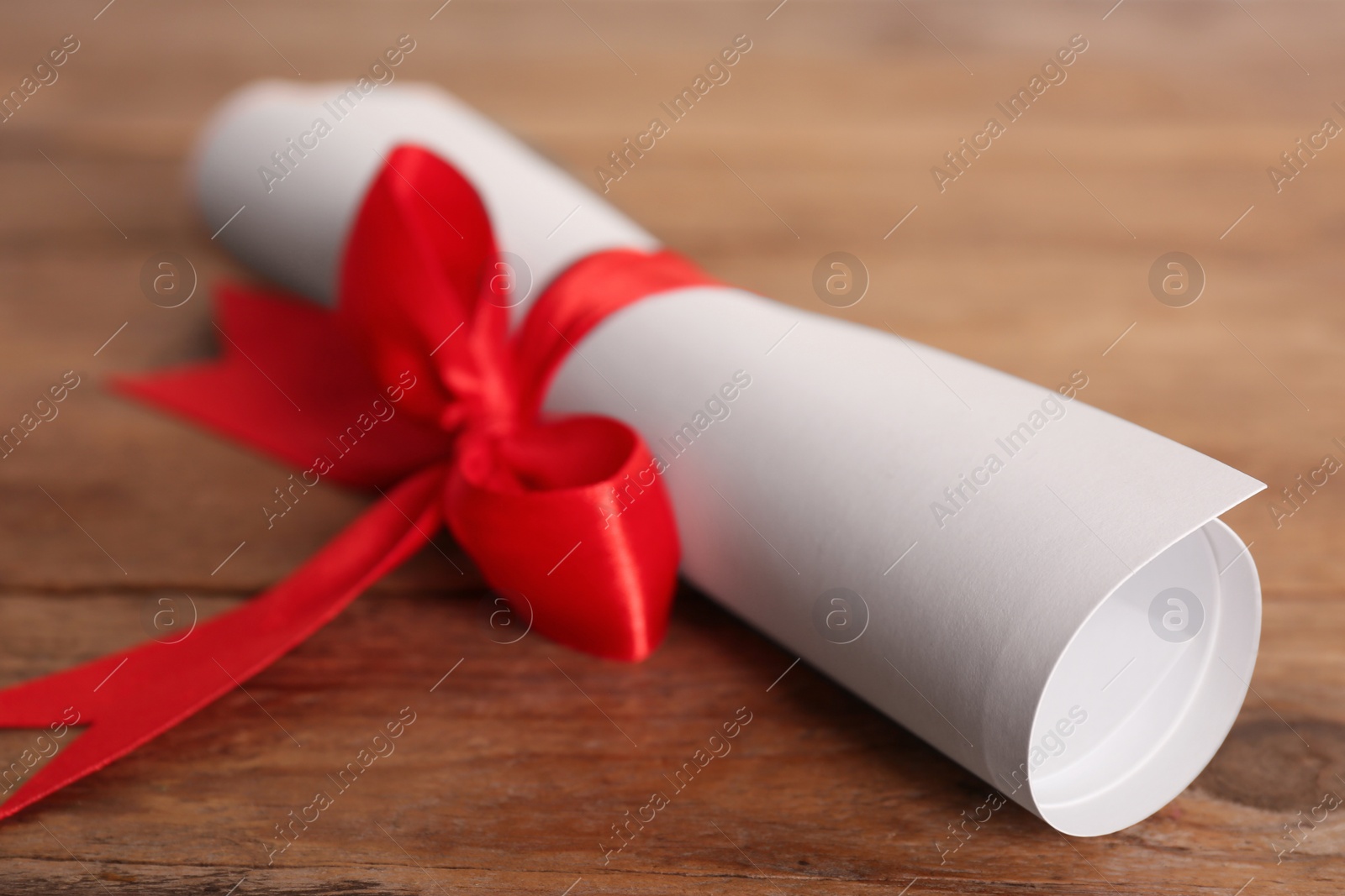 Photo of Rolled student's diploma with red ribbon on wooden table, closeup