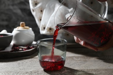 Woman pouring freshly brewed hibiscus tea from teapot into glass at wooden table, closeup