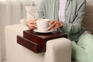 Woman holding cup of drink on sofa armrest wooden table at home, closeup